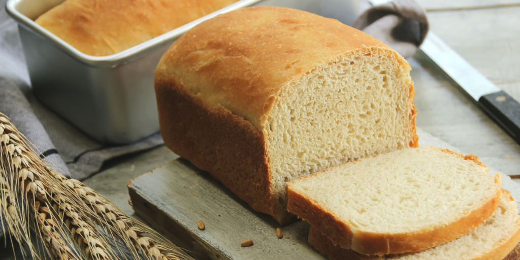 Loaf of bread with sliced bread, wheat grain, loaf pan, and bread knife on cutting board